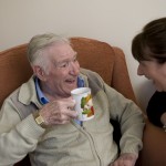 An older man and his Friends of the Elderly Home Support Assistant laughing over tea