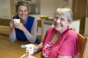 Care worker joins an an elderly woman for a cup of tea while sitting at a kitchen table.