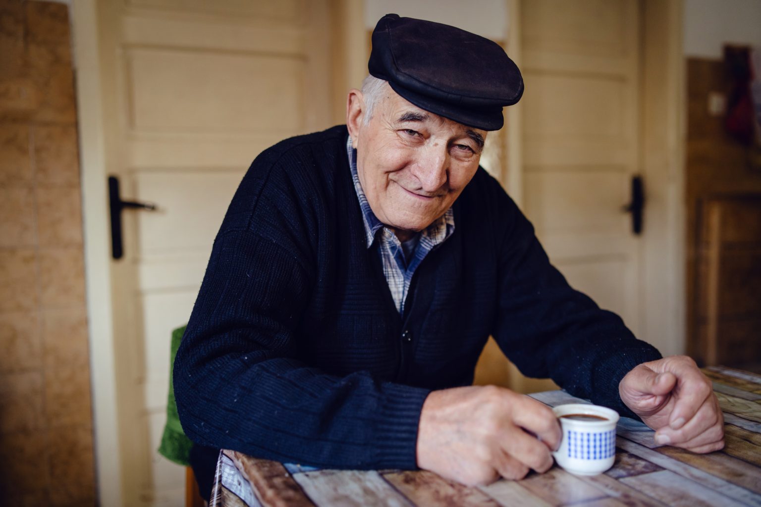 Senior man grandfather old pensioner farmer wearing black sweater and hat having a cup of coffee or tea by the table at home sitting alone smiling