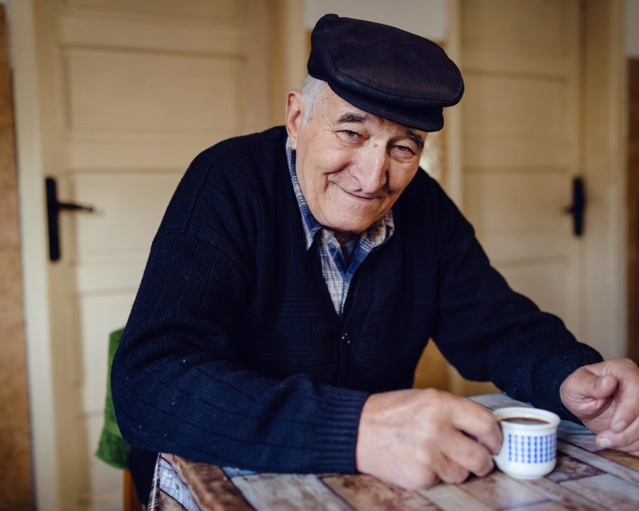 Senior man grandfather old pensioner farmer wearing black sweater and hat having a cup of coffee or tea by the table at home sitting alone smiling