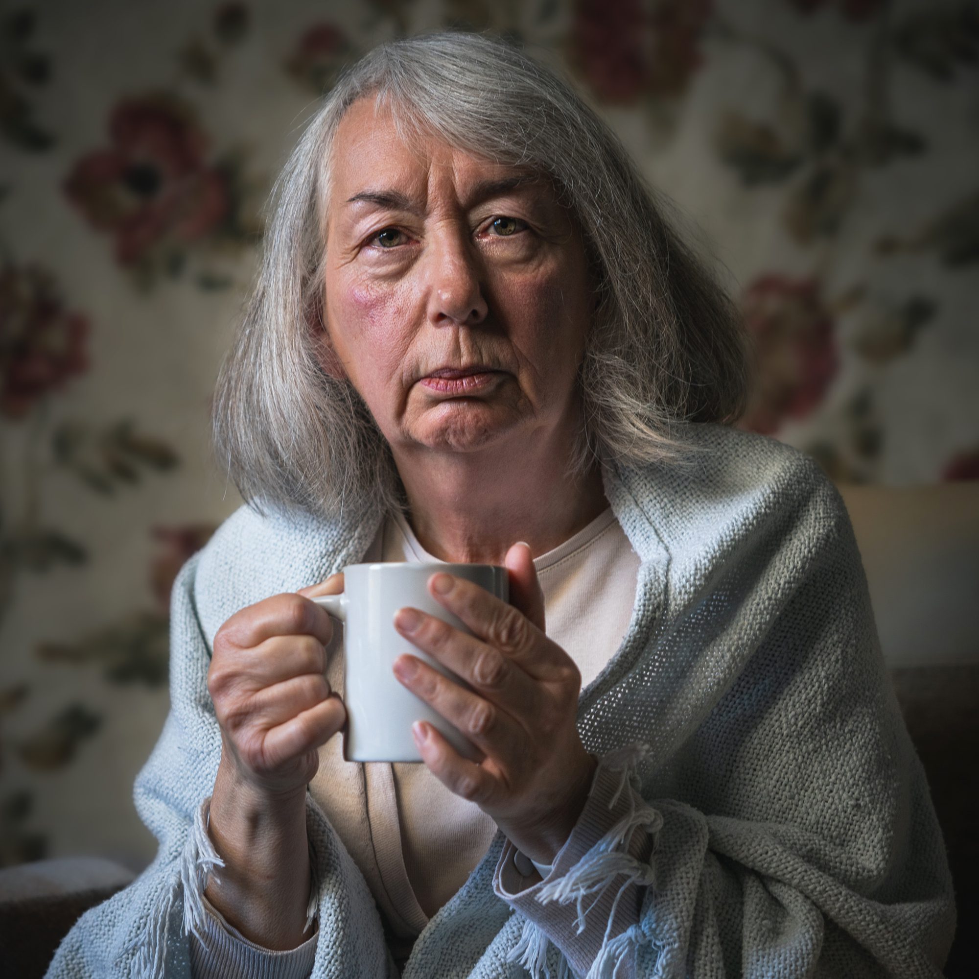 An older woman holding a mug with a blanket around her shoulders.