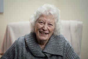 An older woman wearing a grey jumper smiles warmly at the camera.