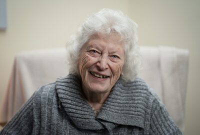 An older woman wearing a grey jumper smiles warmly at the camera.