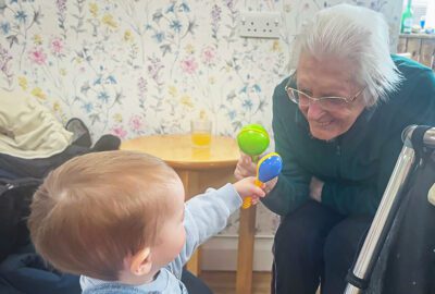 A resident and toddler play with toys together. The resident has a big smile on her face.